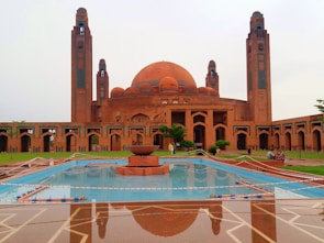 A grand, ornate mosque featuring a large central dome flanked by two tall minarets, with intricate architectural details. The foreground includes a reflective pool with a central fountain and lush green landscaping. The structure is primarily made of reddish-brown brickwork with blue tile accents.