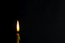 A single candle with a bright flame illuminates a dark, minimalist background. The wax is slightly melted and the flame flickers gently, creating a warm glow.