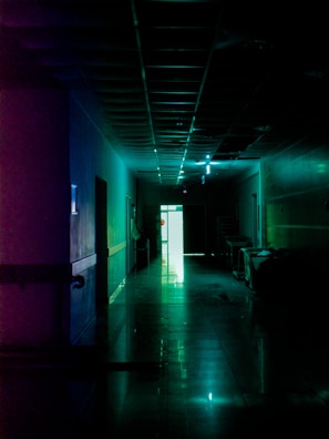 A dimly lit hallway with a polished floor reflecting the neon lights on the ceiling. Some medical equipment is present along the right wall, and the hallway leads to a bright exit at the far end, casting a stark light into the otherwise dark space.