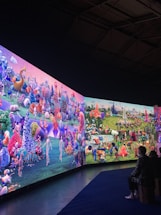 A large, vibrant display featuring a fantastical landscape with diverse, colorful creatures and abstract forms. The scene is filled with imaginative details, including spherical objects, fantastical plants, and humanoid figures. People are seated on benches, observing the immersive digital artworks surrounding them.