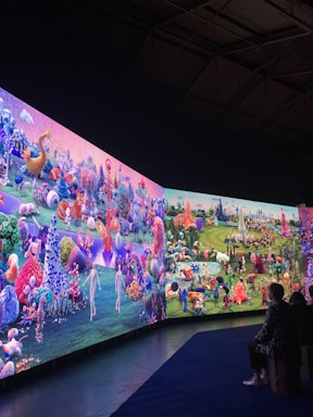 A large, vibrant display featuring a fantastical landscape with diverse, colorful creatures and abstract forms. The scene is filled with imaginative details, including spherical objects, fantastical plants, and humanoid figures. People are seated on benches, observing the immersive digital artworks surrounding them.