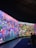 A large, vibrant display featuring a fantastical landscape with diverse, colorful creatures and abstract forms. The scene is filled with imaginative details, including spherical objects, fantastical plants, and humanoid figures. People are seated on benches, observing the immersive digital artworks surrounding them.