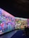 A large, vibrant display featuring a fantastical landscape with diverse, colorful creatures and abstract forms. The scene is filled with imaginative details, including spherical objects, fantastical plants, and humanoid figures. People are seated on benches, observing the immersive digital artworks surrounding them.