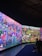 A large, vibrant display featuring a fantastical landscape with diverse, colorful creatures and abstract forms. The scene is filled with imaginative details, including spherical objects, fantastical plants, and humanoid figures. People are seated on benches, observing the immersive digital artworks surrounding them.