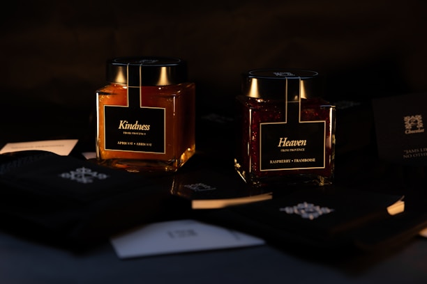 Two elegantly designed jars labeled 'Kindness' and 'Heaven,' containing apricot and raspberry products respectively, are placed against a dark background. The lighting highlights the rich colors of the contents, providing a luxurious and sophisticated atmosphere. Around the jars are various black and white cards with intricate designs.