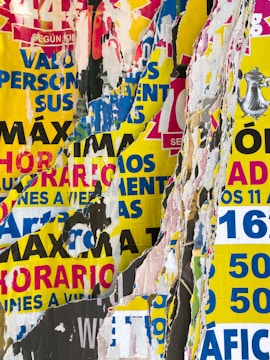 A collage of torn and overlapping colorful posters with various texts in bold colors such as red, blue, yellow, and black. The layers of paper create a textured and chaotic visual effect, with bits of printed words and graphics visible amidst the torn edges.
