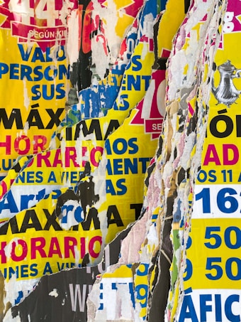 A collage of torn and overlapping colorful posters with various texts in bold colors such as red, blue, yellow, and black. The layers of paper create a textured and chaotic visual effect, with bits of printed words and graphics visible amidst the torn edges.