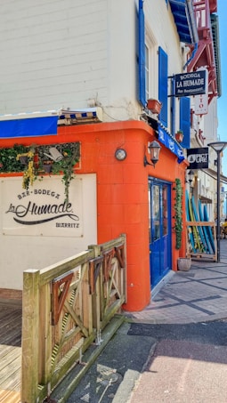 A vibrant street corner featuring a bar and restaurant with rustic wooden accents and a bright orange wall. Blue doors and shutters complement the building, adding a coastal vibe. Surfboards are visible leaning against the wall, and decorative greenery enhances the welcoming atmosphere.