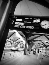 Passengers walk along a modern train platform featuring sleek, high-speed trains. An electronic board overhead displays train information with destinations like Madinah. The architecture is open with arches and large skylights.