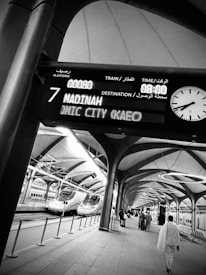 Passengers walk along a modern train platform featuring sleek, high-speed trains. An electronic board overhead displays train information with destinations like Madinah. The architecture is open with arches and large skylights.