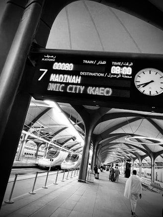 Passengers walk along a modern train platform featuring sleek, high-speed trains. An electronic board overhead displays train information with destinations like Madinah. The architecture is open with arches and large skylights.