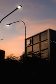 A building with a modern architecture stands with a logo near the top, silhouetted against a warm sunset sky. Streetlights with a soft glow curve over the scene, and dark, leafy trees create a shadowy foreground.