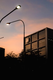 A building with a modern architecture stands with a logo near the top, silhouetted against a warm sunset sky. Streetlights with a soft glow curve over the scene, and dark, leafy trees create a shadowy foreground.