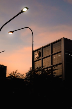 A building with a modern architecture stands with a logo near the top, silhouetted against a warm sunset sky. Streetlights with a soft glow curve over the scene, and dark, leafy trees create a shadowy foreground.