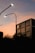 A building with a modern architecture stands with a logo near the top, silhouetted against a warm sunset sky. Streetlights with a soft glow curve over the scene, and dark, leafy trees create a shadowy foreground.