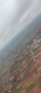 An aerial view of a landscape featuring a mix of agricultural fields with varying shades of green and brown, a network of roads, scattered buildings, and patches of vegetation. The sky above is overcast with a layer of clouds.