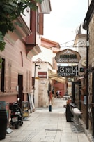 A narrow, cobblestone alley lined with historic buildings featuring rustic architecture. The alley is filled with signs for a lodge hotel, Turkish bath, and rent-a-car services. There are motorbikes parked on the side along with some potted plants, and a few people are walking further down the alley. Bar stools are lined up against one side of a building.