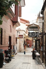 A narrow, cobblestone alley lined with historic buildings featuring rustic architecture. The alley is filled with signs for a lodge hotel, Turkish bath, and rent-a-car services. There are motorbikes parked on the side along with some potted plants, and a few people are walking further down the alley. Bar stools are lined up against one side of a building.
