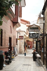 A narrow, cobblestone alley lined with historic buildings featuring rustic architecture. The alley is filled with signs for a lodge hotel, Turkish bath, and rent-a-car services. There are motorbikes parked on the side along with some potted plants, and a few people are walking further down the alley. Bar stools are lined up against one side of a building.