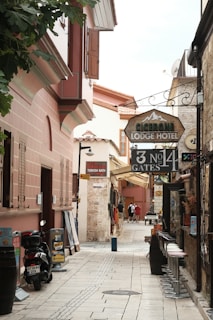 A narrow, cobblestone alley lined with historic buildings featuring rustic architecture. The alley is filled with signs for a lodge hotel, Turkish bath, and rent-a-car services. There are motorbikes parked on the side along with some potted plants, and a few people are walking further down the alley. Bar stools are lined up against one side of a building.