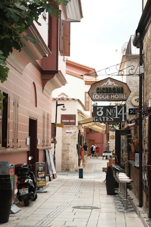 A narrow, cobblestone alley lined with historic buildings featuring rustic architecture. The alley is filled with signs for a lodge hotel, Turkish bath, and rent-a-car services. There are motorbikes parked on the side along with some potted plants, and a few people are walking further down the alley. Bar stools are lined up against one side of a building.
