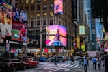 A bustling city street scene with large digital billboards displaying advertisements. Numerous pedestrians and vehicles fill the area, creating a lively urban atmosphere. Bright neon lights and vibrant colors enhance the dynamic environment.