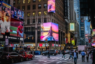 A bustling city street scene with large digital billboards displaying advertisements. Numerous pedestrians and vehicles fill the area, creating a lively urban atmosphere. Bright neon lights and vibrant colors enhance the dynamic environment.