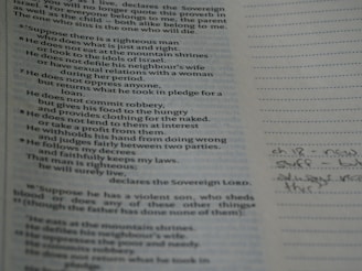 A close-up of a book opened to a page with printed text on the left and lined paper on the right with handwritten notes. The text appears to be religious or scriptural in nature. The page is slightly tilted and the lighting casts a soft shadow.