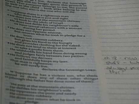 A close-up of a book opened to a page with printed text on the left and lined paper on the right with handwritten notes. The text appears to be religious or scriptural in nature. The page is slightly tilted and the lighting casts a soft shadow.