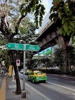 A bustling city street scene featuring a green and yellow taxi on a road lined with trees. Overhead, an elevated train track runs parallel to the street. There is a green directional sign with text in both Thai and English. The atmosphere is urban with a mix of natural greenery, vehicles, and infrastructure.