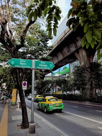 A bustling city street scene featuring a green and yellow taxi on a road lined with trees. Overhead, an elevated train track runs parallel to the street. There is a green directional sign with text in both Thai and English. The atmosphere is urban with a mix of natural greenery, vehicles, and infrastructure.