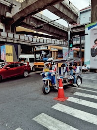 A busy urban street scene featuring a colorful tuk-tuk parked near a pedestrian crossing. Several vehicles, including a bus and a car, are near the tuk-tuk against the backdrop of elevated train tracks. A person is seated inside the tuk-tuk, possibly waiting or using their phone. The area is bustling with signs and traffic cones.