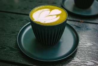 A dark green cup on a matching saucer contains a green matcha latte with a decorative white leaf pattern on top. The setting is a dark wooden table, possibly outdoors.