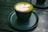 A dark green cup on a matching saucer contains a green matcha latte with a decorative white leaf pattern on top. The setting is a dark wooden table, possibly outdoors.
