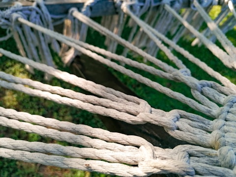Several white ropes are tightly knotted together, creating a pattern with intersecting nodes. The background reveals patches of green grass and a wooden beam, suggesting an outdoor setting.