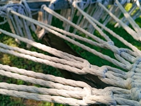 Several white ropes are tightly knotted together, creating a pattern with intersecting nodes. The background reveals patches of green grass and a wooden beam, suggesting an outdoor setting.