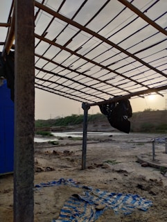 A desolate outdoor setting featuring a makeshift structure with a roof made from wooden slats supported by metal poles. The ground is covered in dirt and debris, including a tattered blue and white striped cloth. In the background, a river flows amidst sandy banks, with sparse vegetation and hills under a hazy sky with the sun low on the horizon.