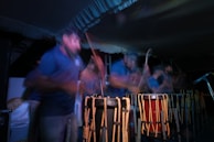 A group of people are engaged in playing traditional drums. The image captures a sense of motion and energy with blurred movements, suggesting an intense and lively performance. The scene is set in low light, highlighting the rhythmic motions and the wooden texture of the drums.