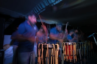 A group of people are engaged in playing traditional drums. The image captures a sense of motion and energy with blurred movements, suggesting an intense and lively performance. The scene is set in low light, highlighting the rhythmic motions and the wooden texture of the drums.