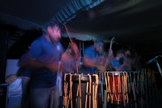 A group of people are engaged in playing traditional drums. The image captures a sense of motion and energy with blurred movements, suggesting an intense and lively performance. The scene is set in low light, highlighting the rhythmic motions and the wooden texture of the drums.