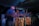 A group of people are engaged in playing traditional drums. The image captures a sense of motion and energy with blurred movements, suggesting an intense and lively performance. The scene is set in low light, highlighting the rhythmic motions and the wooden texture of the drums.