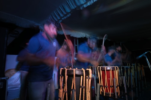 A group of people are engaged in playing traditional drums. The image captures a sense of motion and energy with blurred movements, suggesting an intense and lively performance. The scene is set in low light, highlighting the rhythmic motions and the wooden texture of the drums.