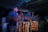 A group of people are engaged in playing traditional drums. The image captures a sense of motion and energy with blurred movements, suggesting an intense and lively performance. The scene is set in low light, highlighting the rhythmic motions and the wooden texture of the drums.