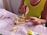 A person wearing a yellow top is sitting at a table, working with playdough. The table is covered with a pink cloth, and various colors of playdough and a stick are visible on the surface. The person is using a pink tool to shape or press into the playdough. The setting suggests a hands-on, creative activity.