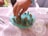 A hand places small natural objects such as cactus spines, pebbles, and leaves onto a light blue modeling clay surface, resting on a table with a decorative tablecloth. Scissors can be seen on the table, indicating a crafting activity.