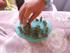 A hand places small natural objects such as cactus spines, pebbles, and leaves onto a light blue modeling clay surface, resting on a table with a decorative tablecloth. Scissors can be seen on the table, indicating a crafting activity.