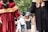 Two people in graduation attire holding hands, walking together in an outdoor setting with blurred individuals in the background. One is wearing a red gown with a decorative sash, the other in a black gown adorned with academic badges.