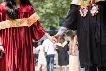 Two people in graduation attire holding hands, walking together in an outdoor setting with blurred individuals in the background. One is wearing a red gown with a decorative sash, the other in a black gown adorned with academic badges.