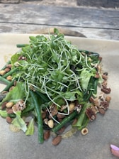 A fresh salad composed of leafy greens, green beans, and a variety of nuts such as almonds and walnuts is arranged on a sheet of parchment paper. The mixture is topped with numerous sprouts, creating a vibrant and rustic display.