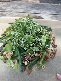 A fresh salad composed of leafy greens, green beans, and a variety of nuts such as almonds and walnuts is arranged on a sheet of parchment paper. The mixture is topped with numerous sprouts, creating a vibrant and rustic display.